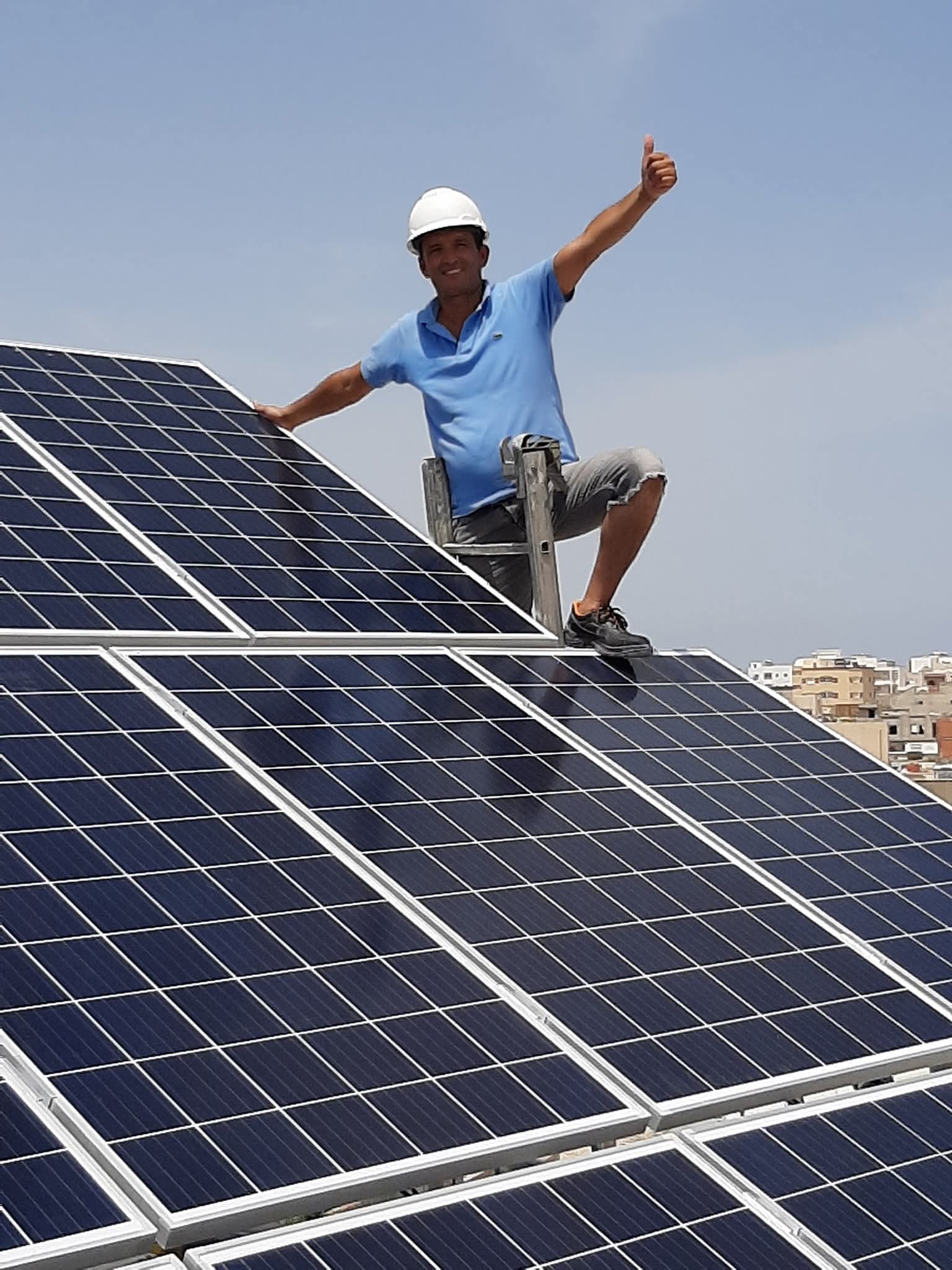 Technicien en casque blanc faisant un pouce levé derrière des panneaux solaires sur un toit.