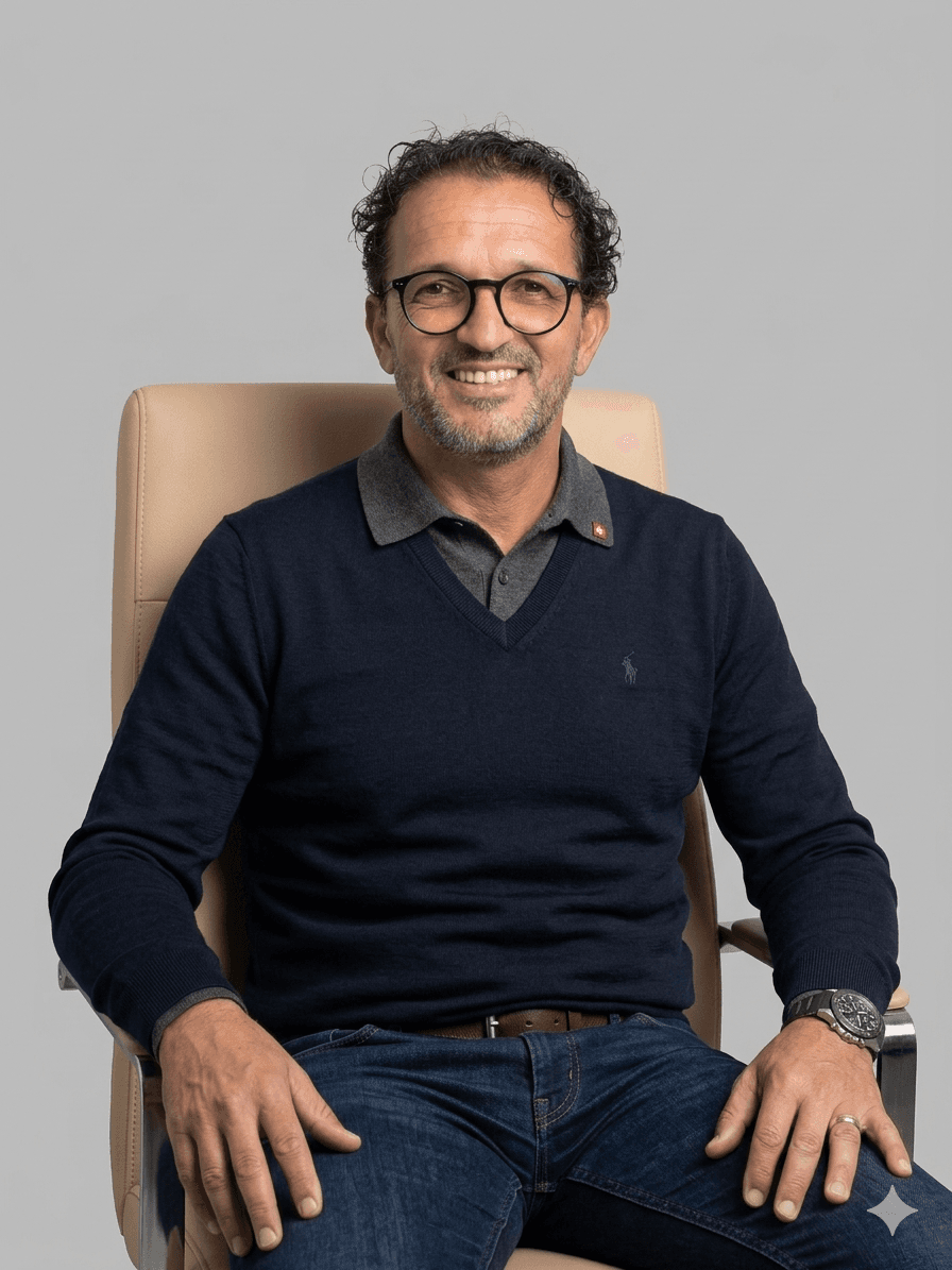 Homme souriant à lunettes, pull bleu marine, assis dans un fauteuil de bureau beige.